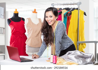 A Portrait Of A Young Asian Designer Woman Using A Laptop And Smiling,clothes Hanged As Background