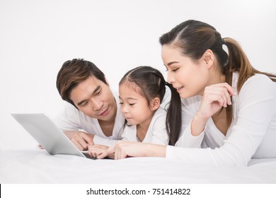 Portrait Of Young Asian Family Father Mother Daughter Look At Tablet Laptop Computer Lying On The Bed, Little Asian Girl Quarantine With Parents. Weekend Holiday Leisure Time Education, Home School.