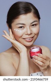 Portrait Of Young Asian Female With Facial Cream Jar And Eye Cream On Face Isolated On Blue Background.