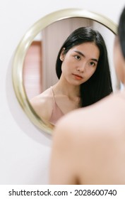 Portrait Of Young Asian Girl With Fresh Healthy Skin Looking At Self In Mirror, Enjoys Youth And Hydration. Natural Makeup Touching Face. Skincare Treatment Cosmetics Concept.