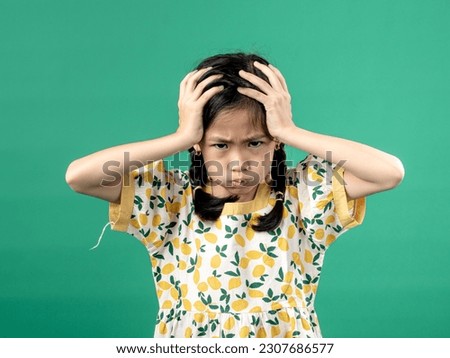 A portrait of a young Asian girl wearing a fruit-patterned dress, seen with a headache as she holds her head and puffs out her cheeks, isolated on a green background.