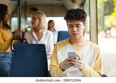 Portrait of young Asian guy traveling on public transit and using cellphone sitting on seat, reading sms message looking at mobile phone screen, chatting online during trip ride inside crowded vehicle