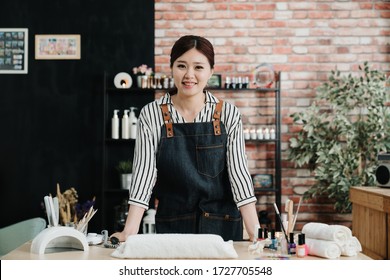 Portrait Of Young Asian Korean Girl Owner Of Beauty Salon For Nail Care And Hands Standing In Studio. Attractive Woman Beautician Face Camera Smiling At Working Table. Manicurist In Apron In Workshop