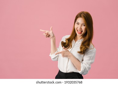 Portrait of young Asian lady smiling with cheerful expression, shows something amazing at blank space in casual clothing and looking at camera isolated over pink background. Facial expression concept.