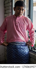 A Portrait Of Young Asian Malay Man Wearing A Pink Color Of Traditional Baju Melayu Cloth With Blue Sampin In The Morning Of Eid Mubarak Or Hari Raya Celebration. Selective Focus.