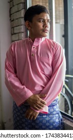 A Portrait Of Young Asian Malay Man Wearing A Pink Color Of Traditional Baju Melayu Cloth With Blue Sampin In The Morning Of Eid Mubarak Or Hari Raya Celebration. Selective Focus.