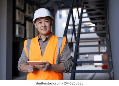 Portrait of a young Asian male builder, engineer and architect standing in a hard hat and vest outside a building, holding a tablet and smiling at the camera.
