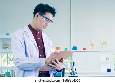 Portrait Of Young Asian Male Scientist Recording Results At Note While Working In The Modern Laboratory Room. The Education Chemistry And Medical Science Research Concept.