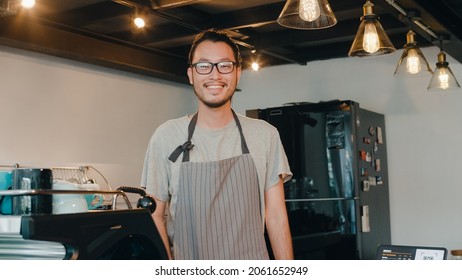 Portrait Young Asian Man Barista Feeling Happy Smiling At Urban Cafe. Small Business Owner Japanese Male In Apron Relax Toothy Smile Looking To Camera Standing At The Counter In Coffee Shop.