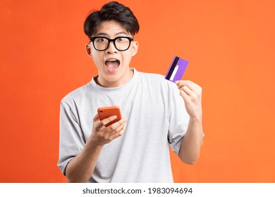 Portrait Of Young Asian Man Holding Smart Phone And ATM Card, Isolated On Orange Background
