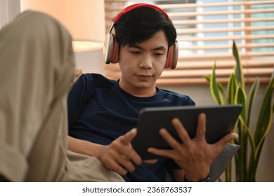 Portrait with Young Asian man listening music on headphones while using digital tablet at home.. - Powered by Shutterstock