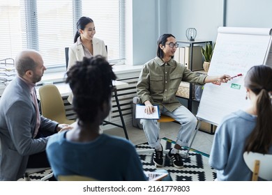 Portrait Of Young Asian Man Pointing At Whiteboard During English Lesson With Diverse Group Of People Sitting In Circle In Office Setting