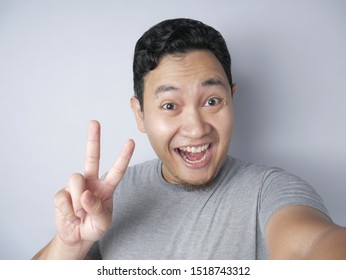 Portrait Of Young Asian Man Smiling While Taking Selfie Photograph Of Himself On His Smart Phone With Funny Expression