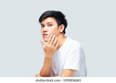 Portrait of a young Asian man wearing a white t-shirt using his hand checking his face on isolated background. Skincare and beauty concept.