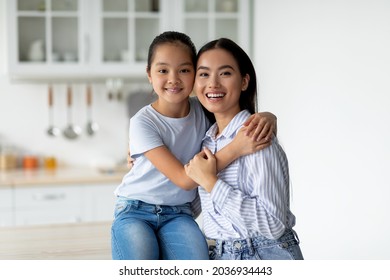 Portrait Of Young Asian Mother And Cute Little Daughter Posing In Kitchen Interior, Pretty Girl Hugging Her Mom And Smiling At Camera, Mommy And Her Child Having Fun At Home