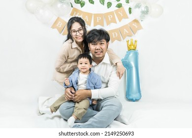 Portrait Of Young Asian Parents Keep In Arms, Hugging Child Kid Baby Boy,celebrating First Birthday Holiday Party On A Minimal Background With Air Balloons.Happy Family:mother,father,son.