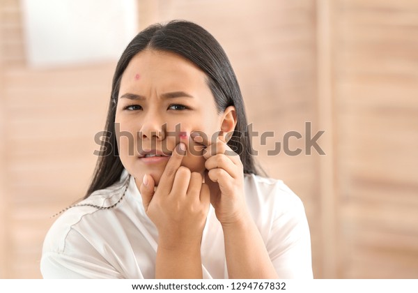Portrait of young Asian woman with acne problem\
squishing pimples in\
bathroom