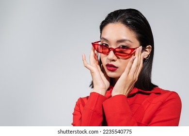 portrait of young asian woman adjusting red and stylish sunglasses while looking at camera isolated on grey