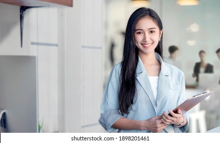 Portrait of young asian woman in casual wear holdng clipboard, smiling and looking at camera while standing in modern office room. - Powered by Shutterstock