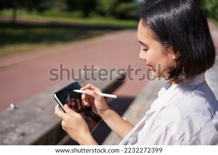 Portrait of young asian woman drawing on fresh air in park, sitting with graphic tablet and digital pen, smiling happy.