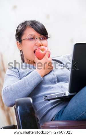 Portrait of a young Asian woman eating apple while looking at her laptop on sofa.