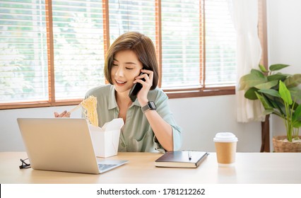 Portrait Of Young Asian Woman Enjoying Chinese Food In Office Lunch Break, Female Employee Holding Chopsticks Eating Takeaway Noodle Box Meal At Workplace In Home Office. 