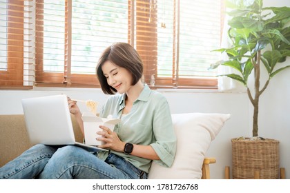 Portrait Of Young Asian Woman Enjoying Chinese Food In Living Room Lunch Break. Asia Girl Holding Chopsticks Eating Takeaway Noodle Box Meal In Home Office. Social Distancing Quarantine Virtual.