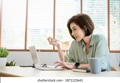 Portrait Of Young Asian Woman Enjoying Chinese Food In Office Lunch Break, Female Employee Holding Chopsticks Eating Takeaway Noodle Box Meal At Workplace In Home Office. 