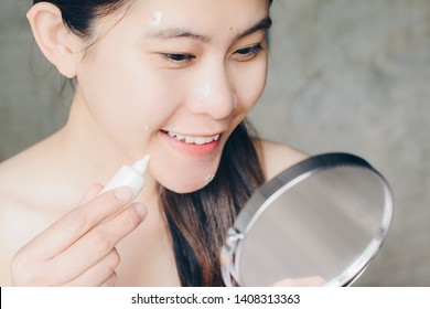 Portrait Of Young Asian Woman Holding Cosmetic Tube And Applying Acne Cream Or Moisturizer On Her Face By Looking A Pocket Mirror. Conceptual Of Beauty And Cosmetic.