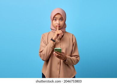 Portrait Of Young Asian Woman Holding Mobile Phone And Makes Silence Gesture With Fingers