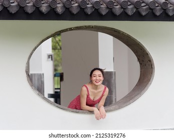 Portrait of young Asian woman looking and smiling from oval window in park, beautiful Chinese girl in sexy red sundress enjoy her carefree time in hot summer day.