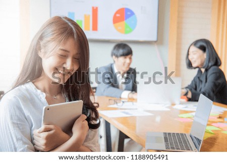Portrait of young asian woman in office with coworkers talking in background. Female creative professional looking at camera and smiling.