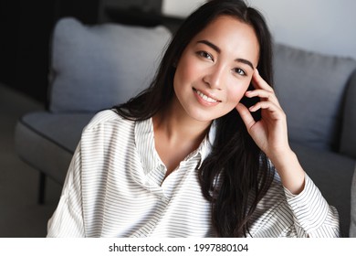 Portrait Of Young Asian Woman Sitting At Home In Living Room, Girl With Glowing Natural Skin, No Make Up, Smiling Happy At Camera, Resting And Relaxing On Weekend Inside House, Leisure Time