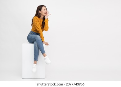Portrait of young Asian woman sitting on white box and keeps hand near mouth and whispers secret isolated over white background, Spreads rumors concept