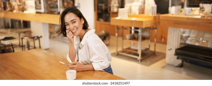 Portrait of young asian woman, sitting in cafe, holding smartphone, chatting and messaging while drinking coffee. - Powered by Shutterstock