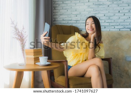 Portrait young asian woman using mobile phone with coffee cup and read book sit on chair in living room interior