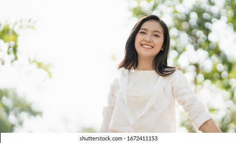 Portrait Of Young Asian Woman Wearing The White Shirt In Casual Style Smiling And Enjoy Life In The Natural Sunlight In The Park, Relaxing Moment, Enjoying The Nature, Mindful And Feel The Fresh Are.