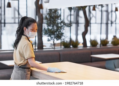 Portrait Of Young Asian Woman Wearing Mask While Cleaning Tables In Modern Restaurant Interior, Covid Safety Concept, Copy Space