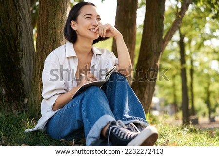 Portrait of young asian woman writing in her notebook, expressing her thoughts on paper in diary, smiling and sitting under tree on summer day in park.