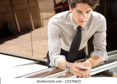 Portrait Of A Young Aspirational Businessman Using A Smart Phone While Leaning On A Banister Outside A Modern Glass Office Building In The City.