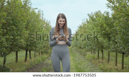 Portrait of a young athletic fitness girl holding a phone in her hands. Shooting on the street on the pyrode between the trees. The girl reads a message from the phone and uses the program, smiling