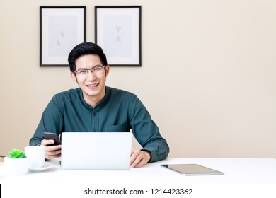 Portrait Of Young Attractive Asian Businessman Or Student Using Mobile Phone, Laptop, Tablet, Drinking Coffee Sitting On Desk Table Wearing Green Shirt Look At Camera In Home Office With Copy Space.