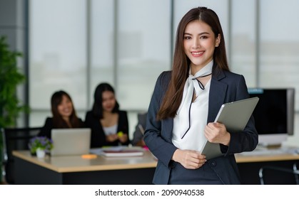 Portrait Of Young Attractive Asian Female Office Worker In Formal Business Suits  Smiling At Camera In Office With Blurry Colleagues Sitting In Office As Background.