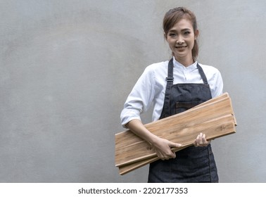 Portrait Young Attractive Asian Female Carpenter Holding Wood Planks In Carpentry Wood Working Workshop, Cheerful Carpenter Woman Wear Apron And Hold Wood Prepare For DIY Wood Craft, Copy Space