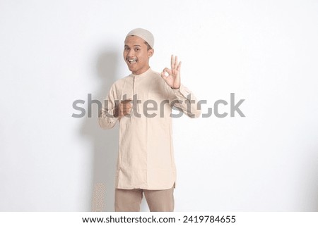 Portrait of young attractive Asian muslim man in koko shirt with skullcap showing ok hand gesture and smiling looking at camera. Approval concept. Isolated image on white background