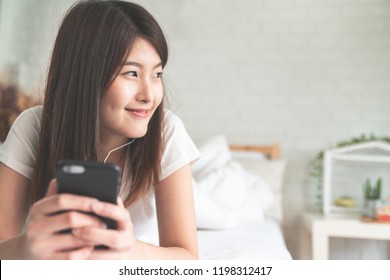 Portrait Of Young Attractive Asian Woman Listening To Music In Smartphone Smiling Relax On White Bed And Cozy Bedroom With Soft Tone. Young Happy Student Or College Female Lifestyle At Home Concept.