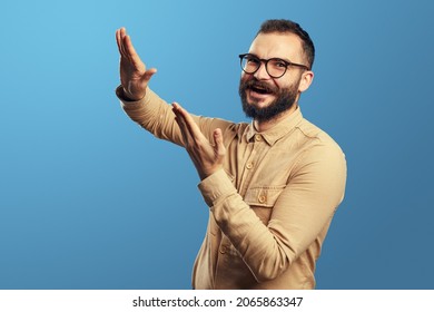 Portrait of young attractive bearded man in beige shirt gesturing throwing away money, having fun in studio, isolated over blue background