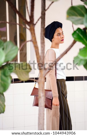 Portrait of a young, attractive, beautiful and elegant Muslim (Arab, Asian, Malay) woman wearing a turban (hijab, head scarf) through several beautiful plants against a plain off white background. 