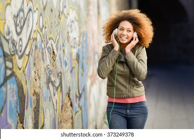 Portrait Of Young Attractive Black Girl In Urban Background Listening To The Music With Headphones. Woman Wearing Leather Jacket And Blue Jeans With Afro Hairstyle