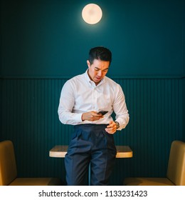 Portrait of young and attractive Chinese Asian business man using his smartphone and smiling. He is professionally dressed in a fitting shirt and pants in the office. 